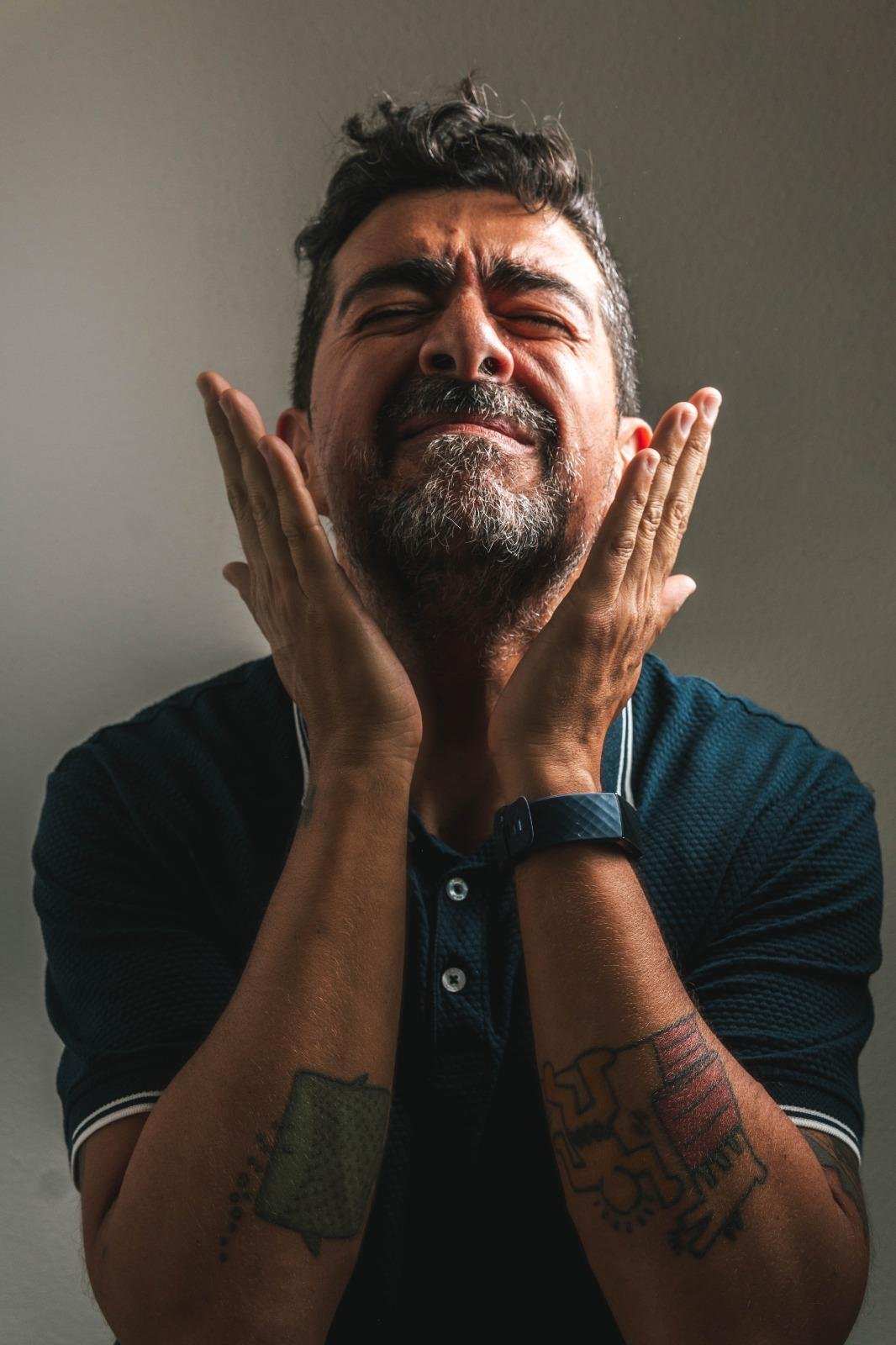 Middle-aged man with beard and dark hair wearing navy blue shirt, hands near face in emotional expression, with tattoos visible on arms